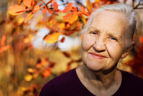 Elderly woman outside smiling with sun shining on her face and fall leaves in the background
