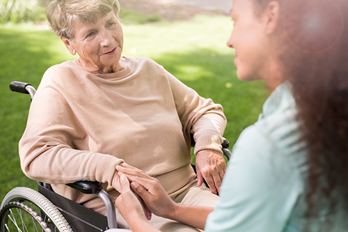 Young nurse in teal scrubs knelt down in front of elderly woman in wheelchair holding her hand